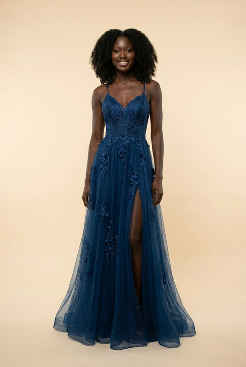 Femme portant une robe de bal évasée en tulle marine, bustier en dentelle, décolleté cœur et fente haute, devant un décor méditerranéen.