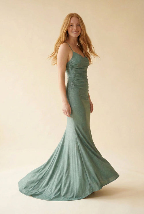 Femme portant une robe de bal sirène vert sauge scintillante à bretelles fines et décolleté en cœur, avec traîne, devant des parois rocheuses.